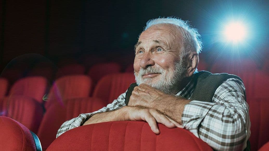 Ein Senior mit grauen Haaren und Bart sitzt in einem Kino und schaut lächelnd einen Film.