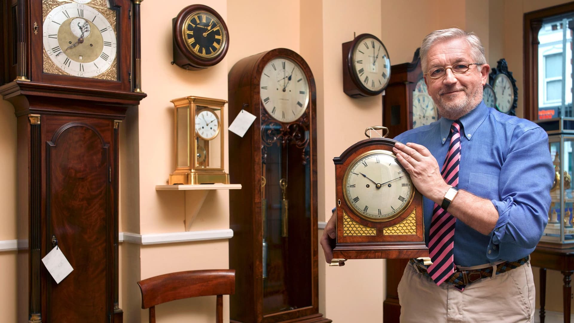 Ein Uhrmacher mit grauen Haaren steht in seinem Laden und hält lächelnd eine alte Schrankuhr in seinen Händen.