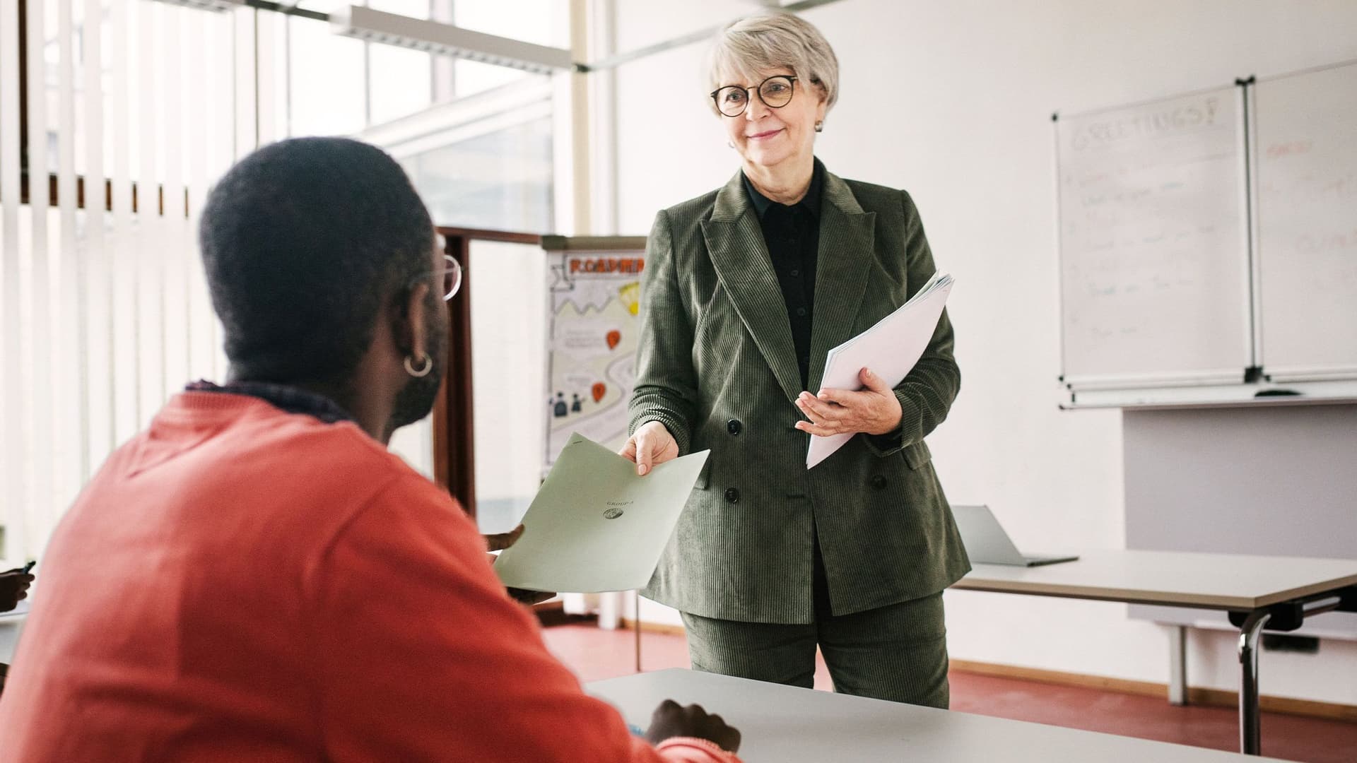 Eine Lehrerin mit grauen Haaren übergibt einem jungen, erwachsenen Schüler Papierpögen in einem Klassenraum.
