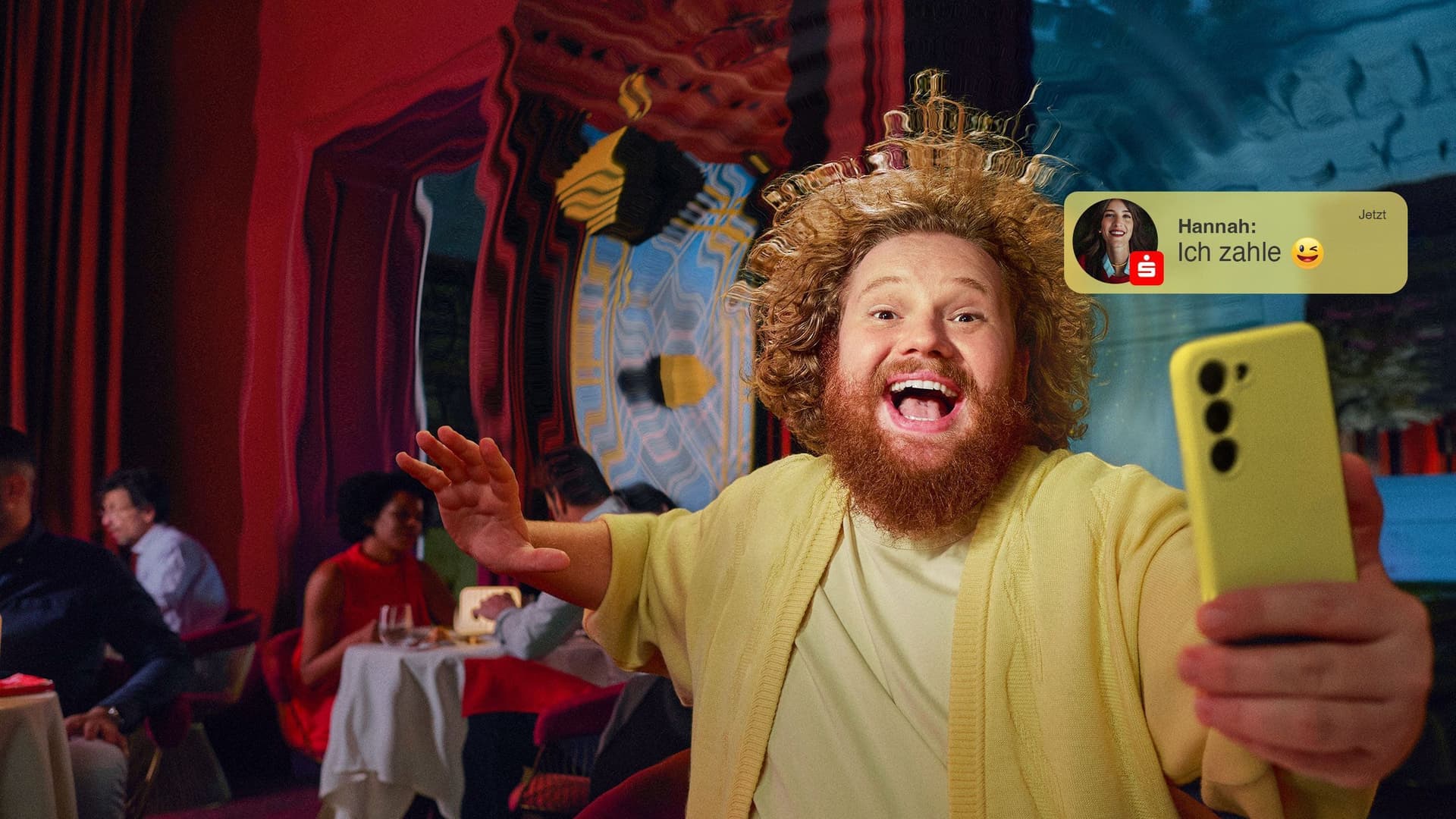 Ein fröhlicher Mann mit lockigem Haar und Bart hält ein gelbes Smartphone in der Hand und lacht in die Kamera. Auf dem Bildschirm erscheint eine Benachrichtigung mit dem Text „Hannah: Ich zahle“ von der Sparkasse-App