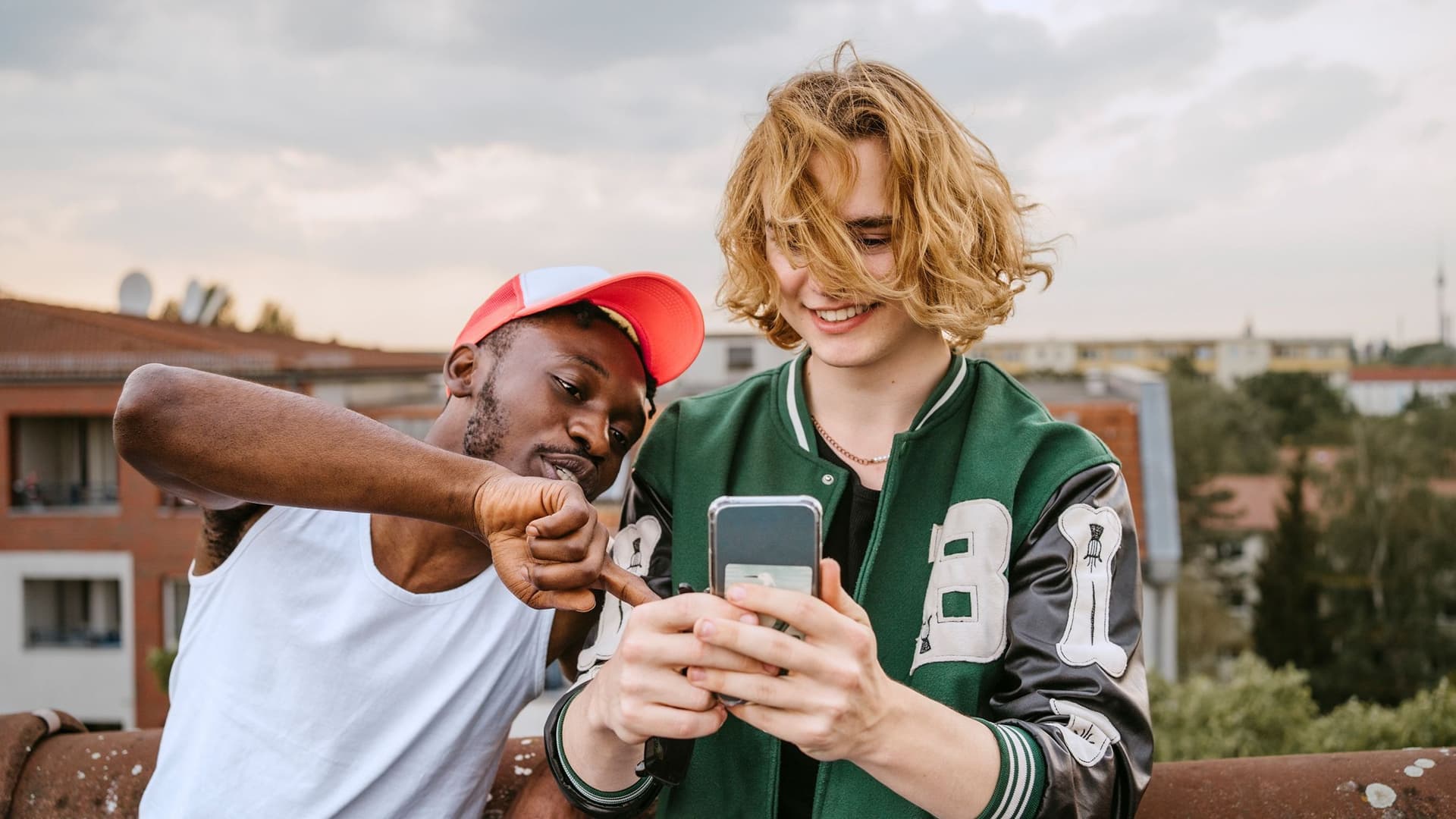 Zwei junge, erwachsene Männer, in sportlich modischer, Freizeitkleidung, stehen auf dem Dach in einer Wohnsiedlung und sehen zusammen auf den Screen eines Smartphones.