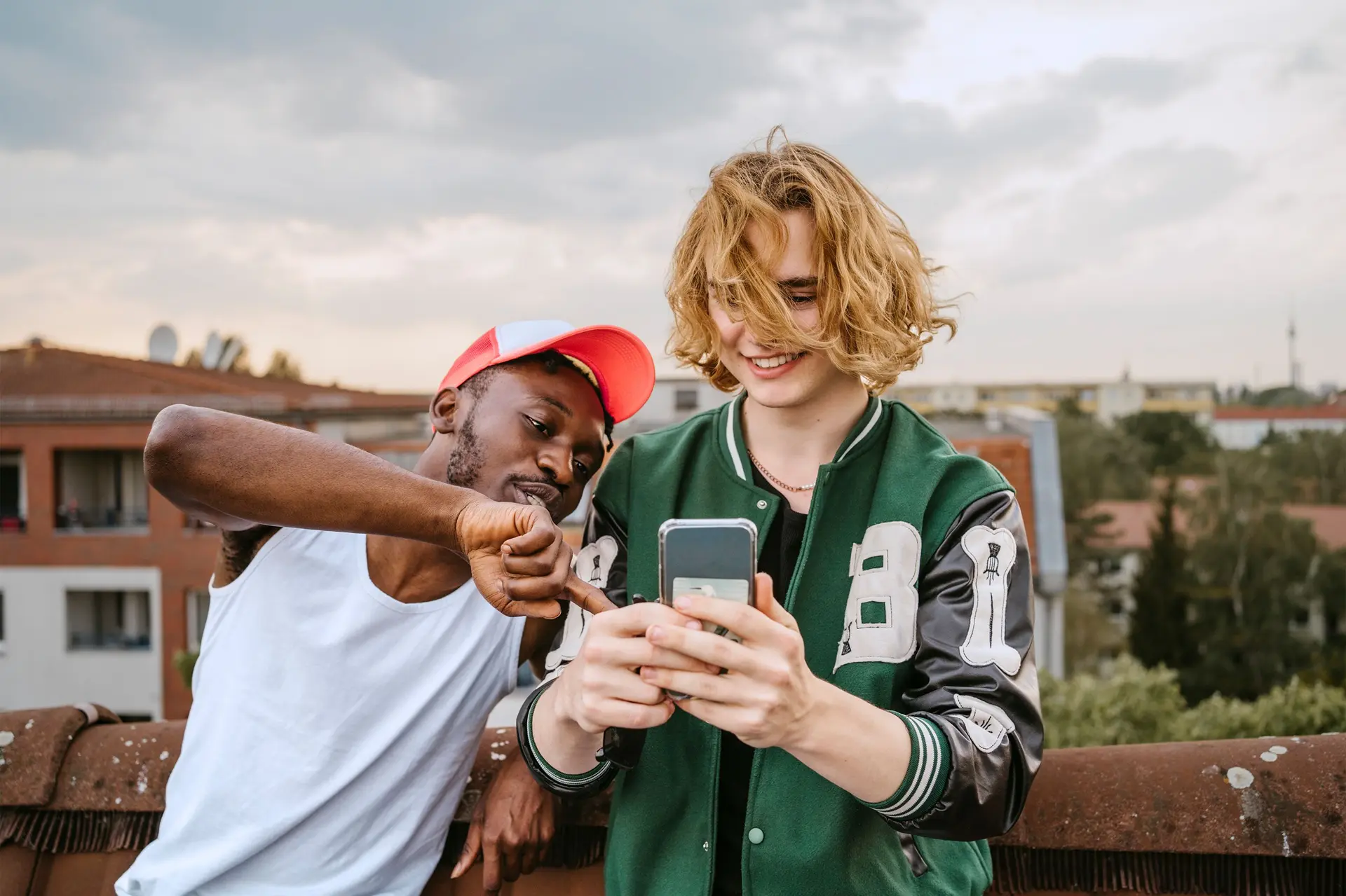 Zwei junge, erwachsene Männer, in sportlich modischer, Freizeitkleidung, stehen auf dem Dach in einer Wohnsiedlung und sehen zusammen auf den Screen eines Smartphones.