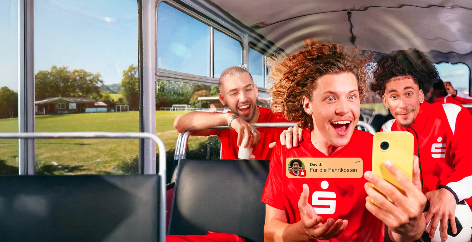 Junge Fußballspieler in roten Trikots sitzen fröhlich in einem Bus; im Vordergrund hält ein Spieler ein gelbes Smartphone, im Bild ist zusätzlich eine Push-Nachricht zu sehen: „David: Für die Fahrtkosten“.