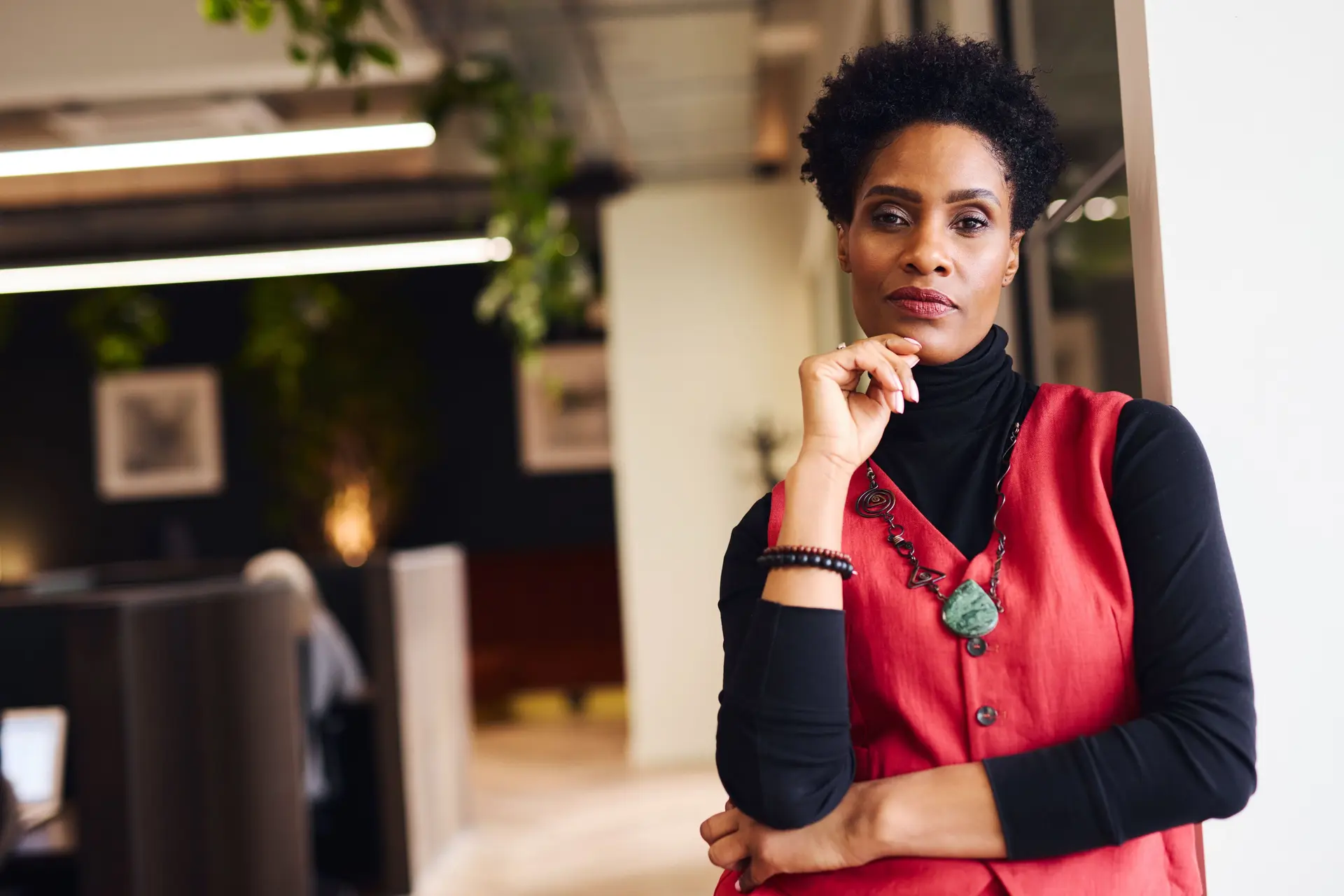Porträt einer mittelalten Frau mit der Hand am Kinn, die mit nachdenklicher Miene im Coworking Space in die Kamera blickt.