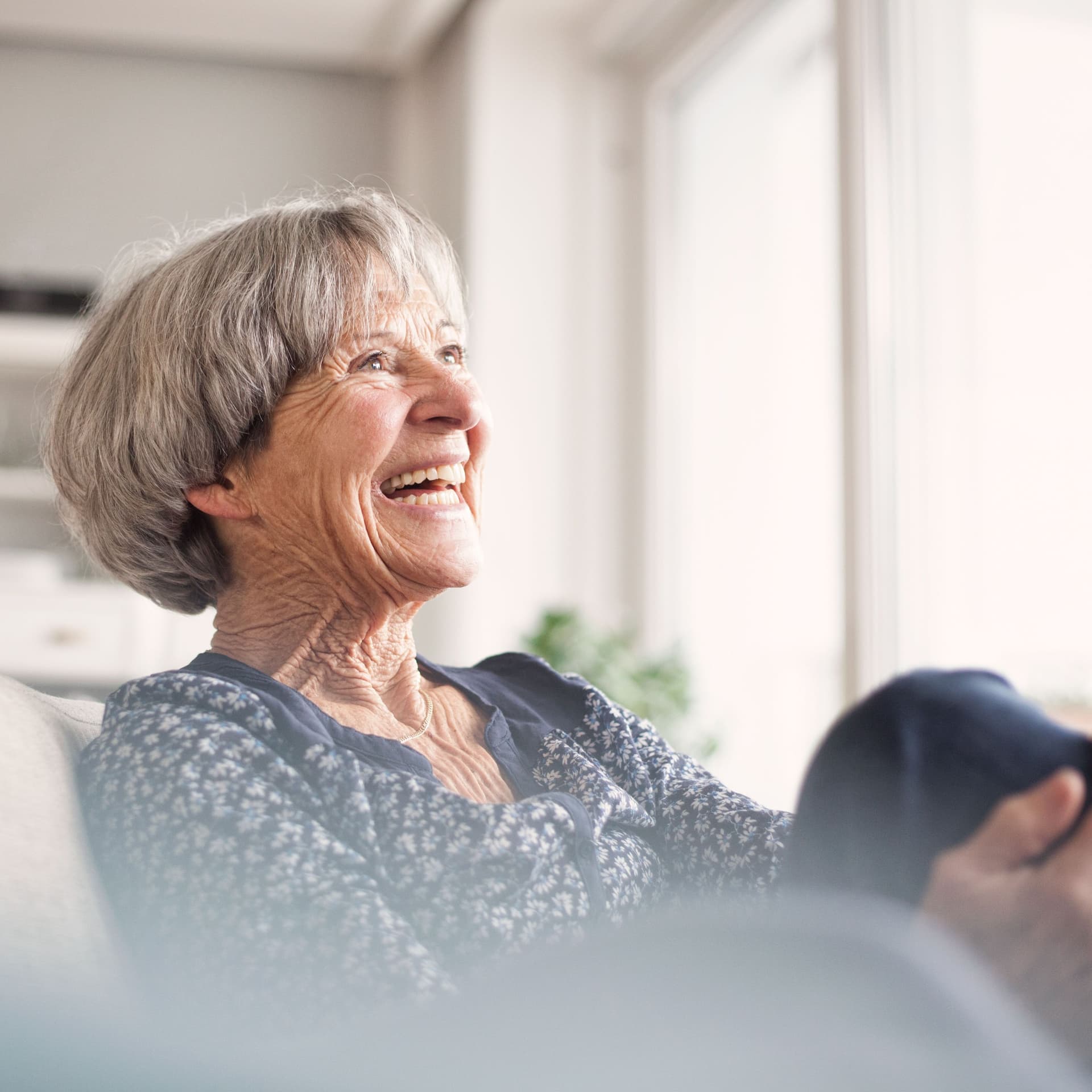 Porträt einer lachenden älteren Frau, die auf einem grauen Sofa sitzt.