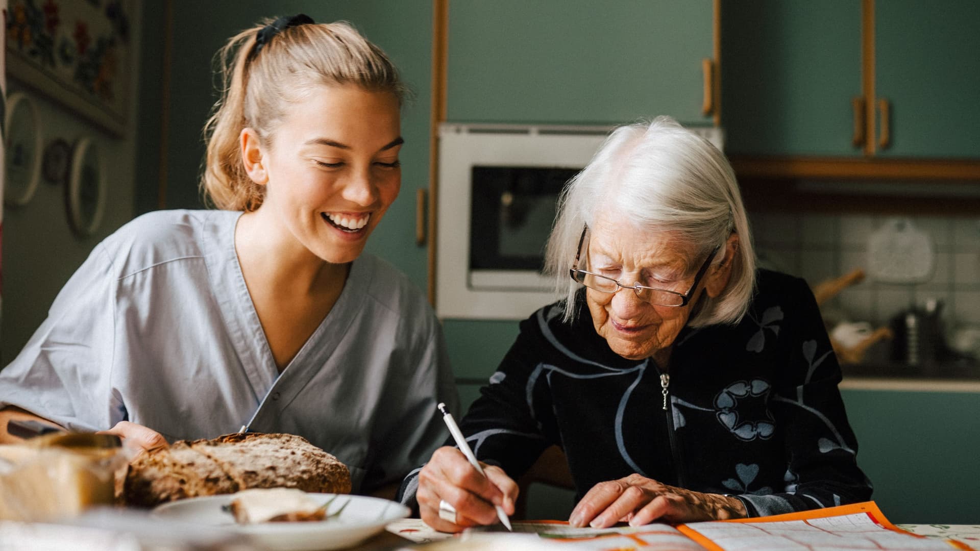 Ältere und jüngere Frau sitzen gemeinsam an gedecktem Küchentisch. Die Ältere hält einen Stift in der Hand und löst ein Kreuzworträtsel.