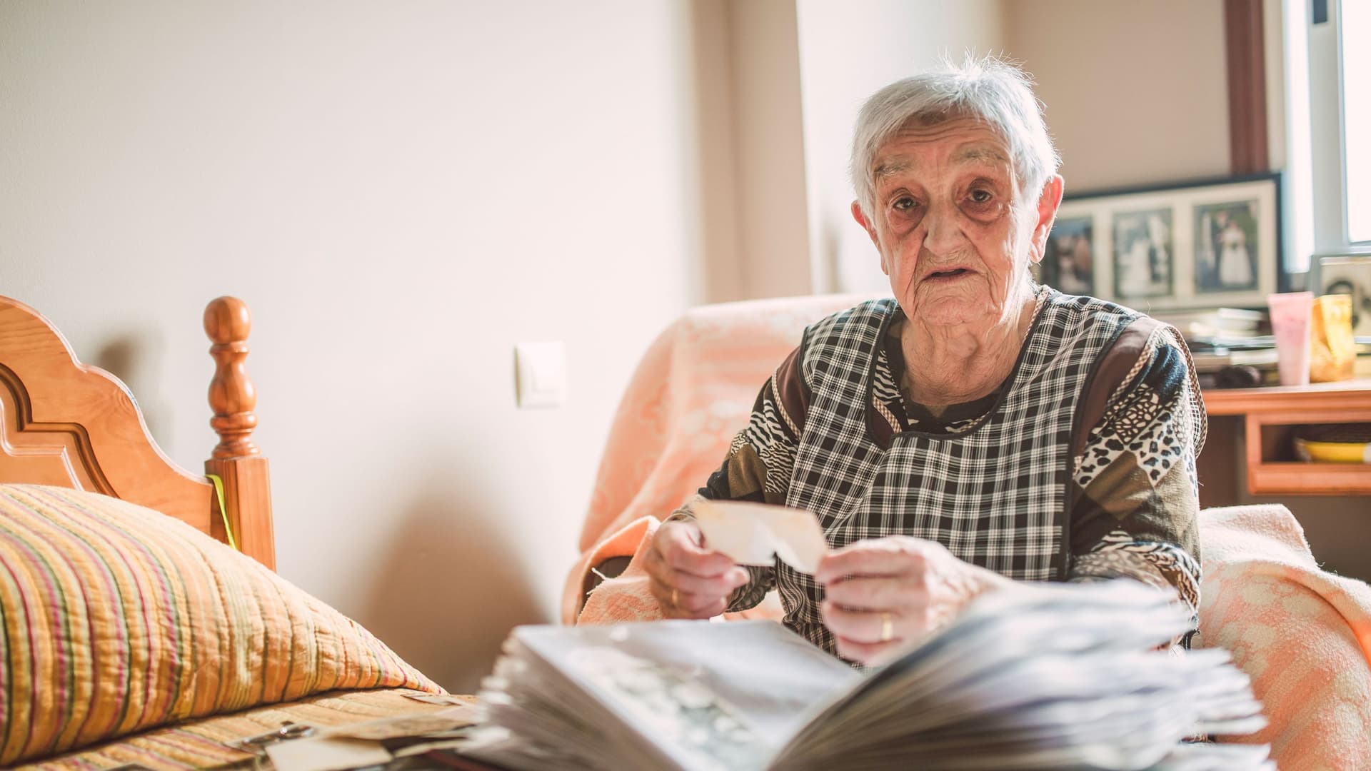Eine Seniorin sitzt mit einem Fotoalbum vor einem Bett und blickt in die Kamera