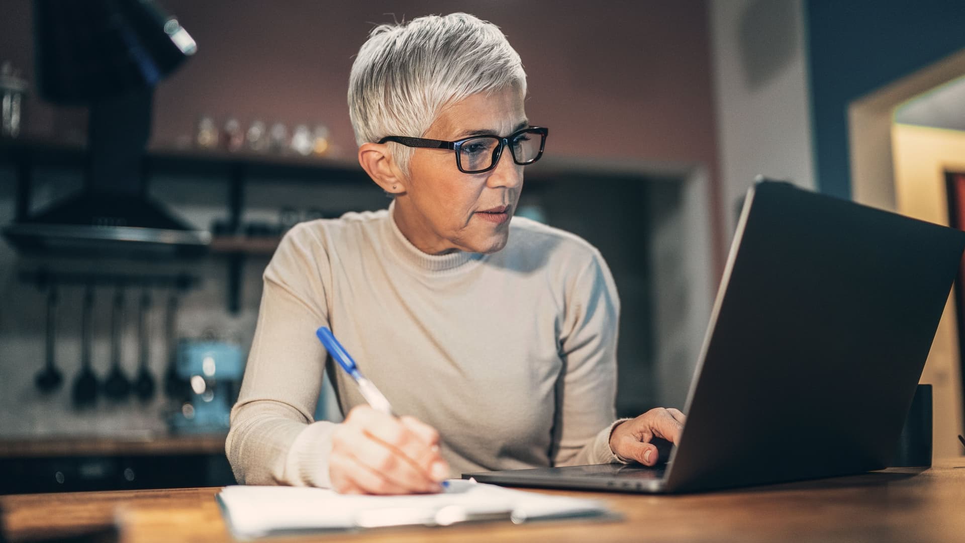 Ältere Frau sitzt vor einem Laptop in der Küche. Sie hält einen Stift in der rechten Hand, Papier liegt auf dem Tisch.
