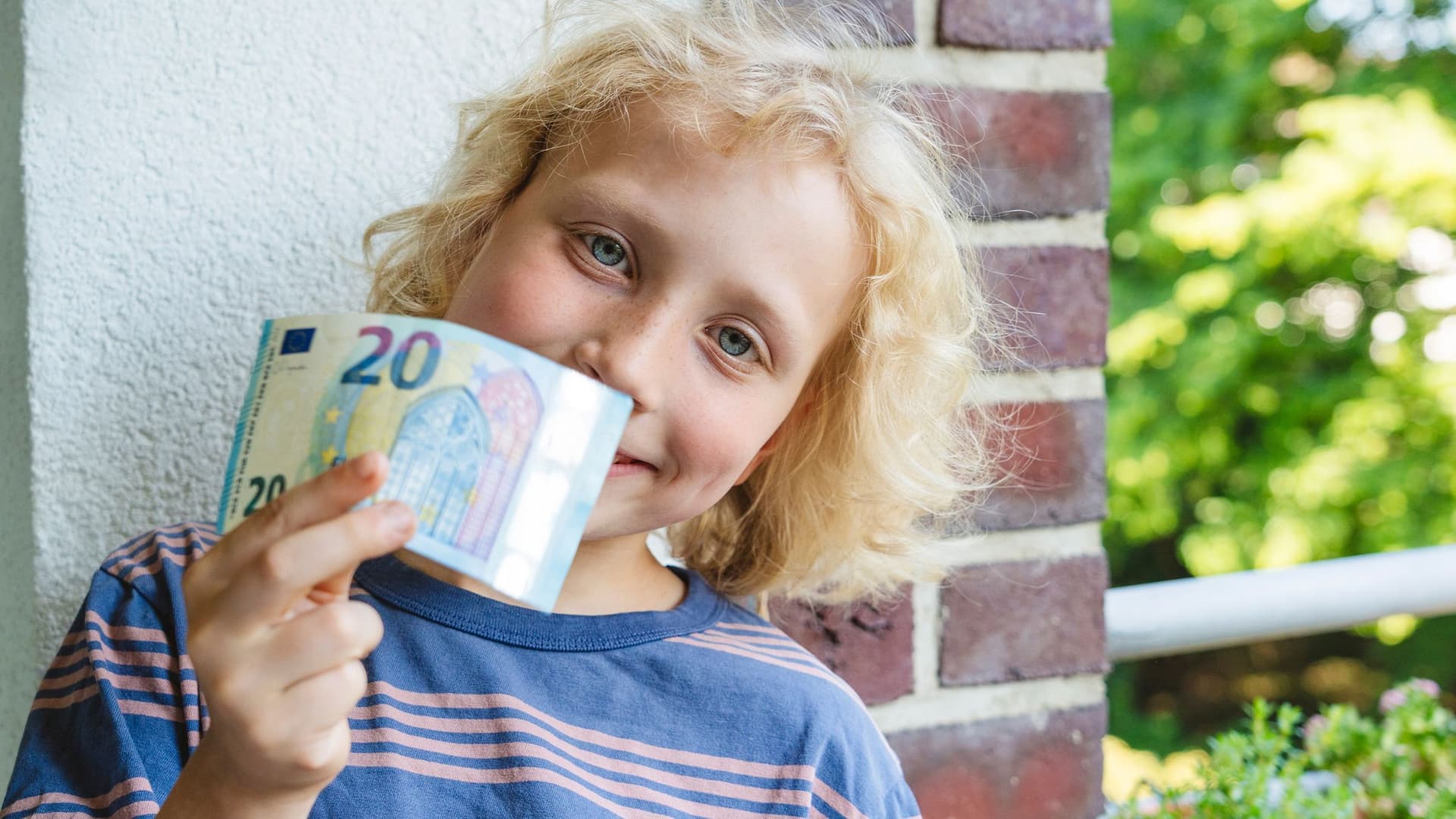 Eine bunte Bildkomposition mit einem Jungen im Grundschulalter, der freudig einen Zwanzig-Euro-Schein in die Kamera hält.