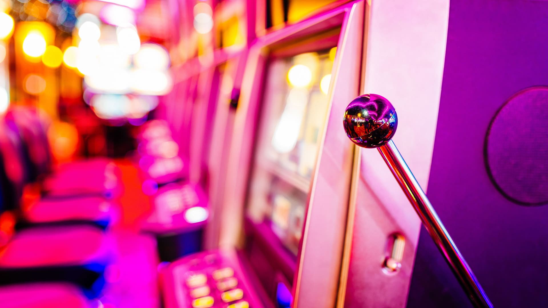 Seitliche Aufnahme auf eine Reihe von Spielautomaten mit Sitzplätzen davor. Sie werden von buntem LED Licht angestrahlt.