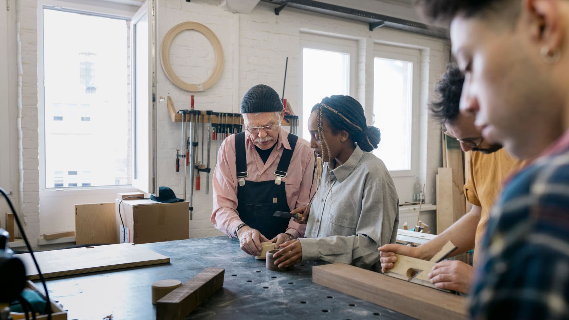 Eine junge Frau und zwei junge Männer arbeiten zusammen mit einem älteren Holzwerkstatttechniker in einer Werkstatt. Sie haben Holzblöcke vor sich.