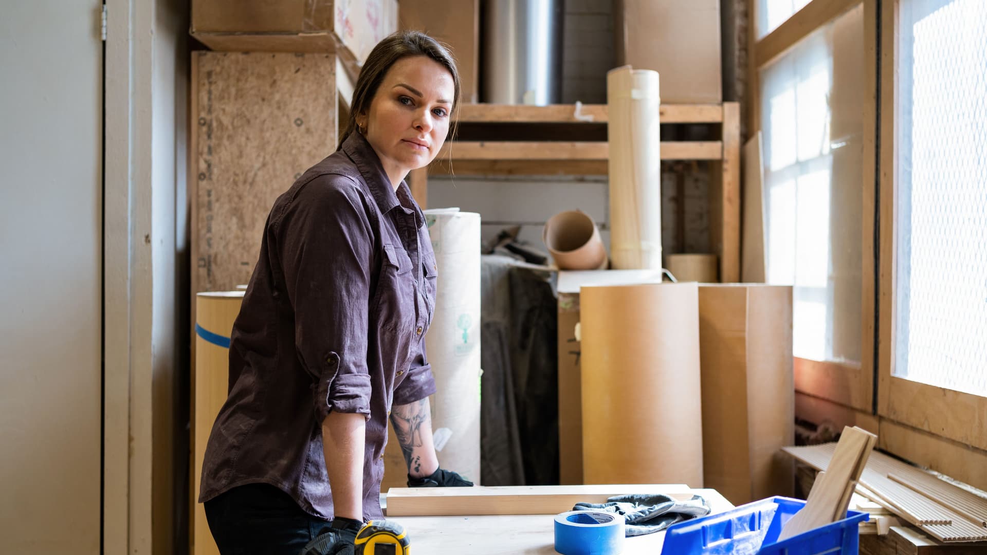 Frau, die an einer Werkbank in einer Werkstatt steht. Sie hat die Hände aufgestützt und guckt in die Kamera.