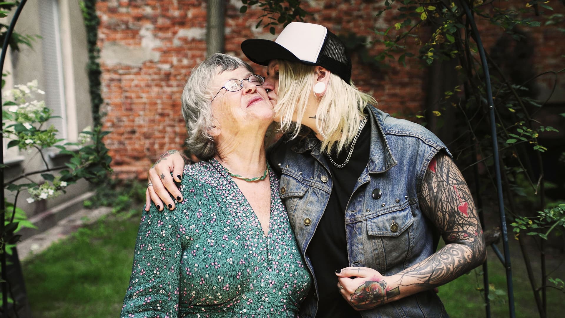 Junge Frau küsst eine ältere Frau auf die Wange und legt den Arm und sie, während sie in einem grünen Hinterhof steht.