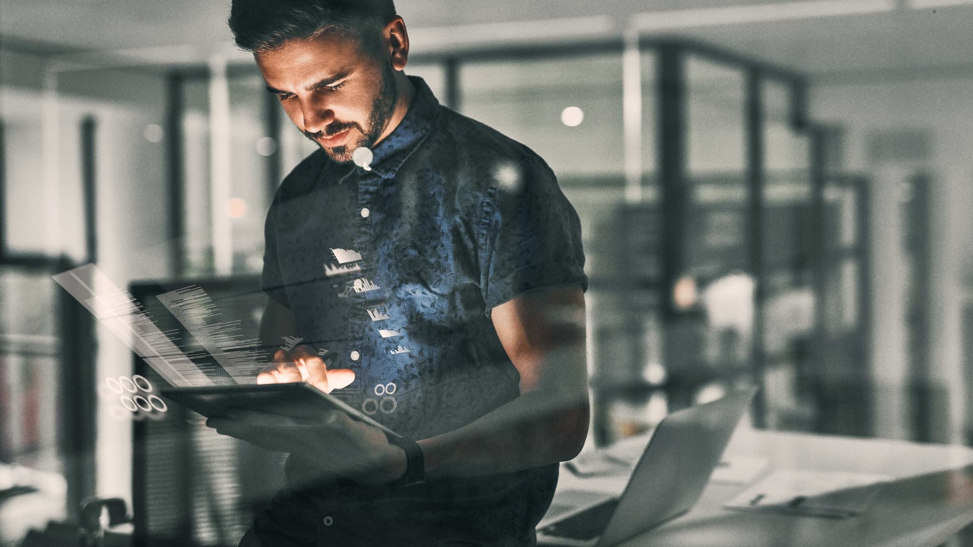 Junger Mann schaut im halbdunklen auf ein Tablet. Das Büro hinter ihm ist leer.