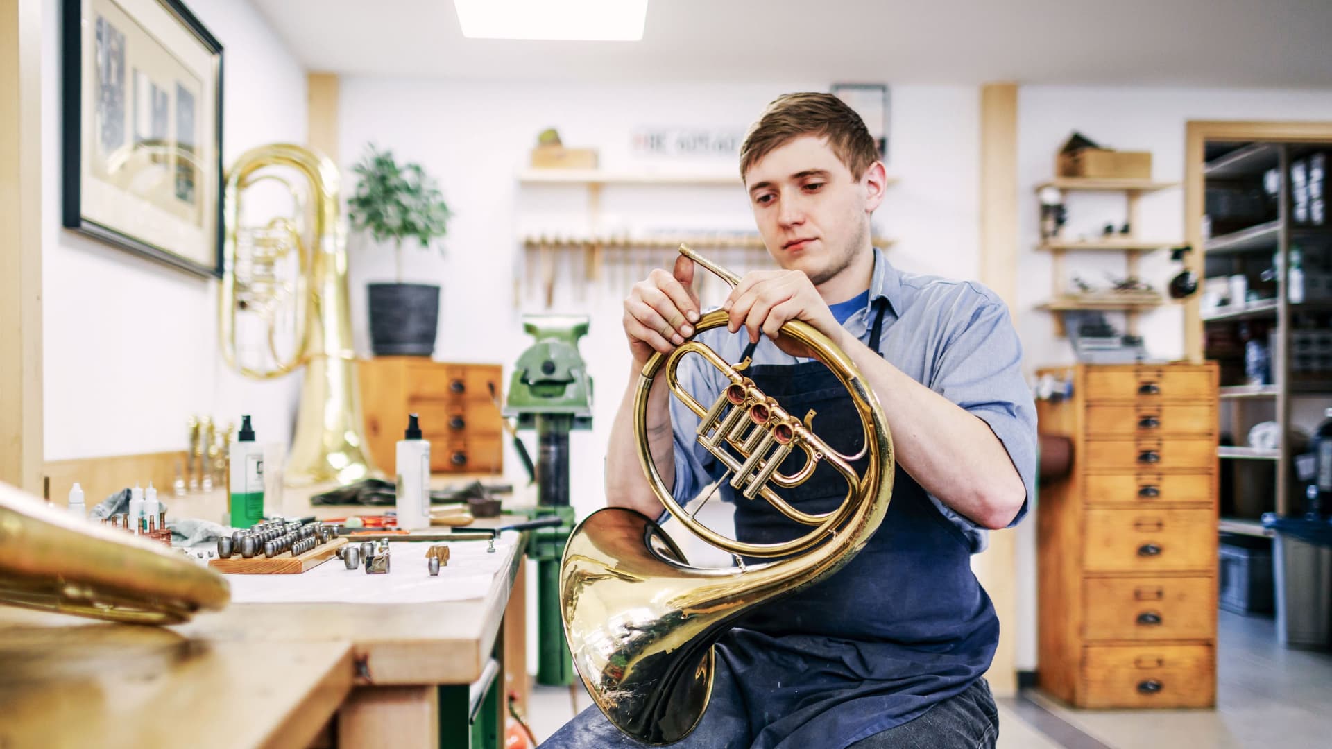 Ein junger Mann sitzt in einer Werkstatt und arbeitet an einem Blasinstrument. Er trägt eine Schürze und ist ganz vertieft.