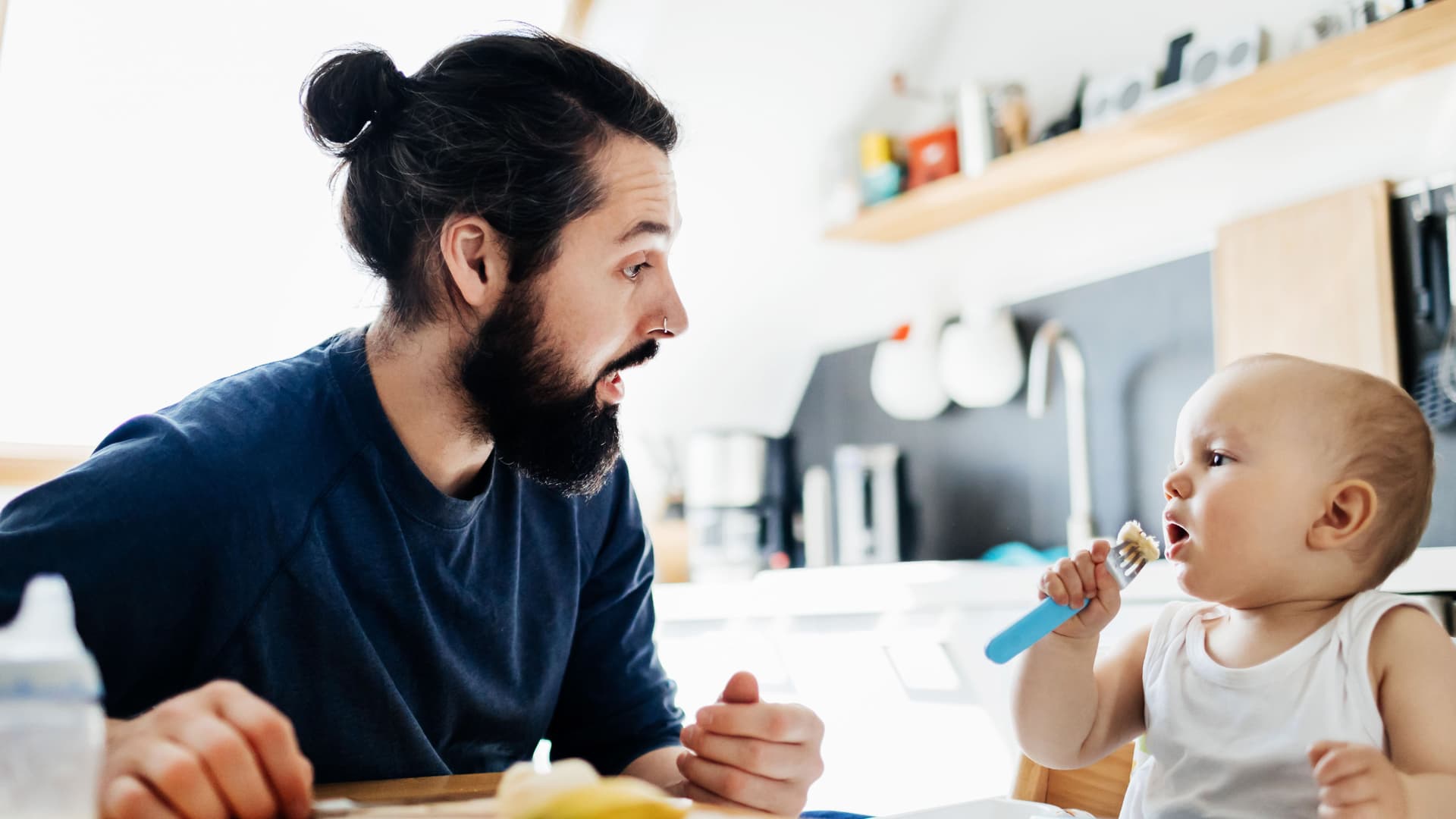 Sparbuch für ein Kind eröffnen Ein junger Mann und ein Baby im Hochstuhl sitzen an einem Küchentisch. Das Baby isst Brei von einem Löffel und der Mann guckt es mit geöffnetem Mund an.