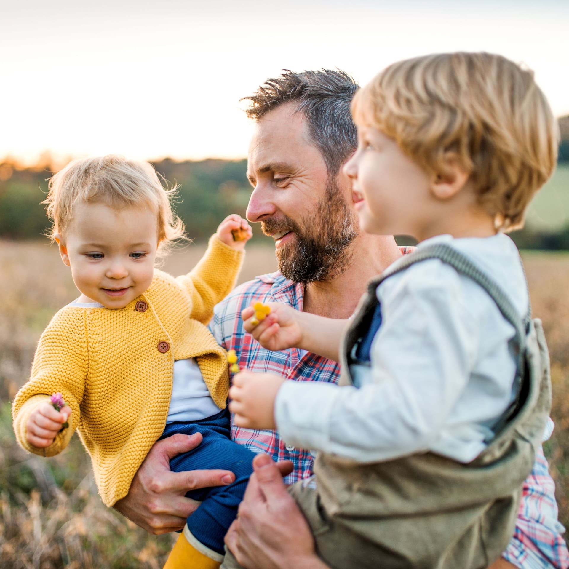Ein Vater mit zwei Kleinkindern auf dem Arm auf einer Herbstwiese im Freien.