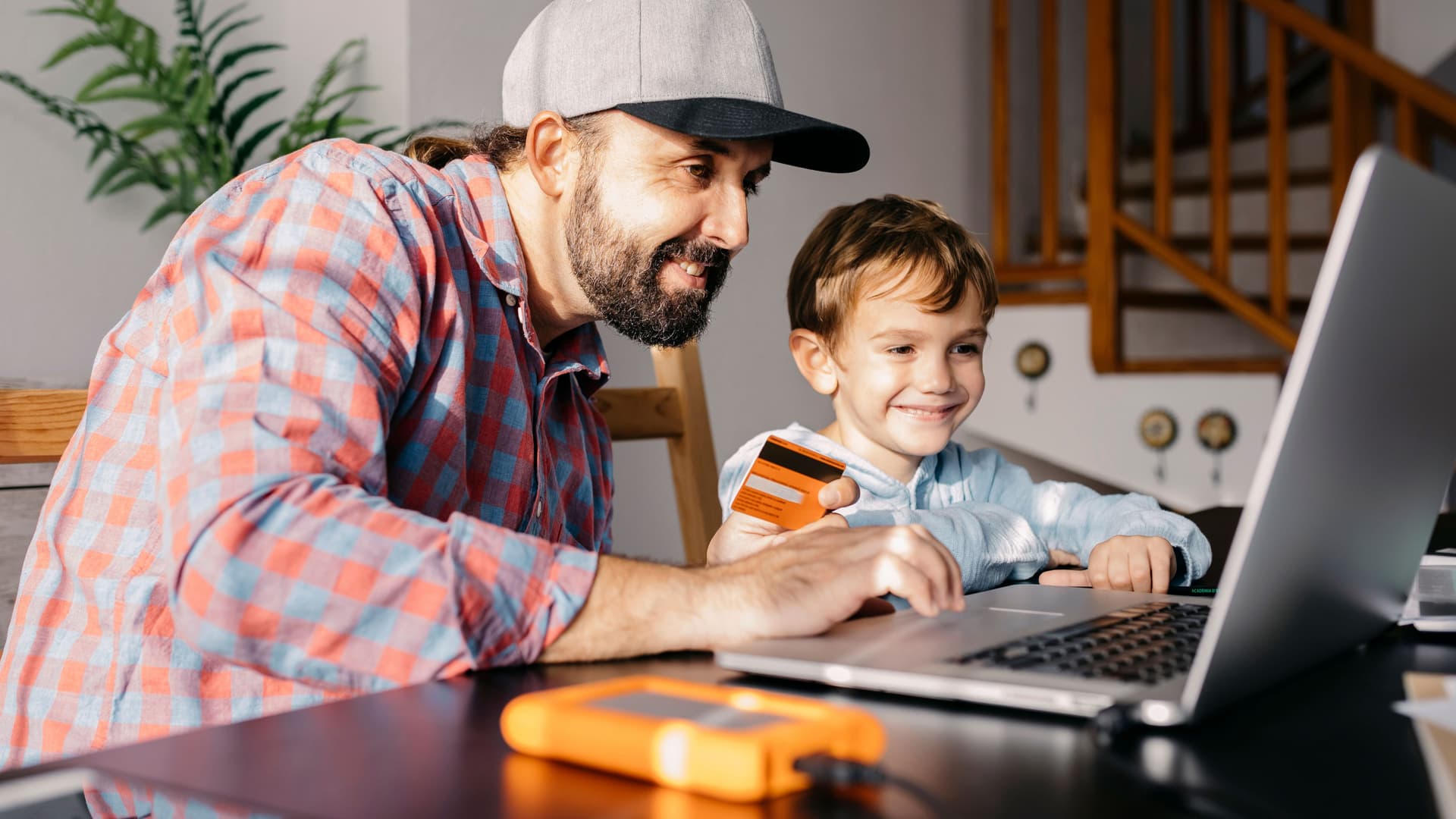 Mann und Kind sitzen am Laptop. Mann hält eine Kreditkarte in der Hand.