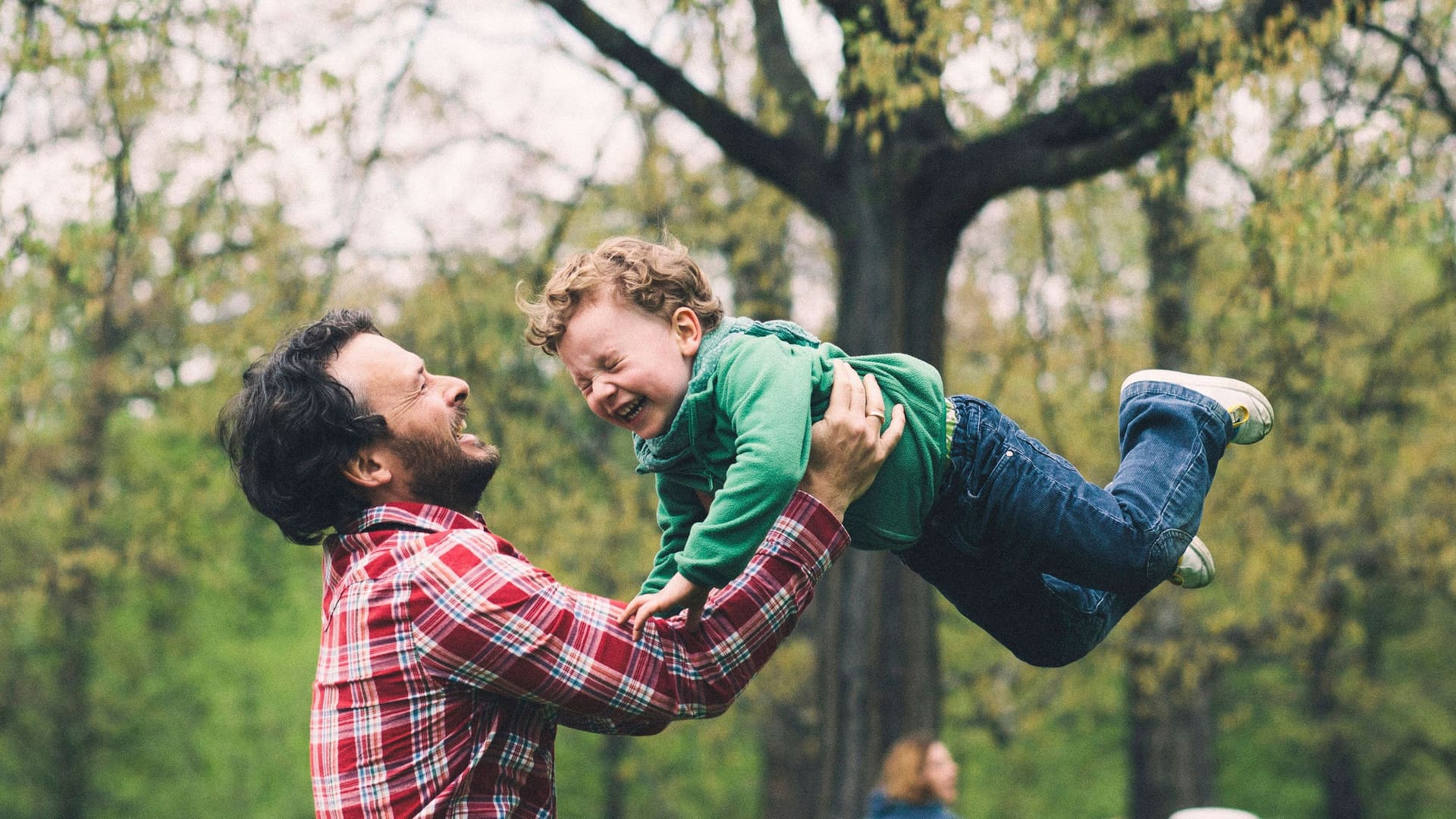 Vater wirbelt seinen Sohn im Park herum. Die Mutter spielt im Hintergrund Frisbee.