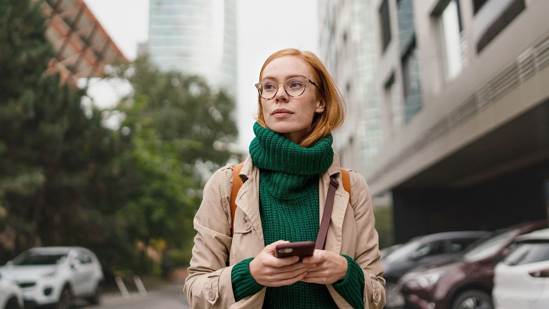 Einer herbstlich gekleidete Frau mit einem Handy in der Hand läuft durch eine Großstadt. Sie schaut nach vorne in die Ferne. Im Hintergrund zeichnen sich Hochhäuser ab.