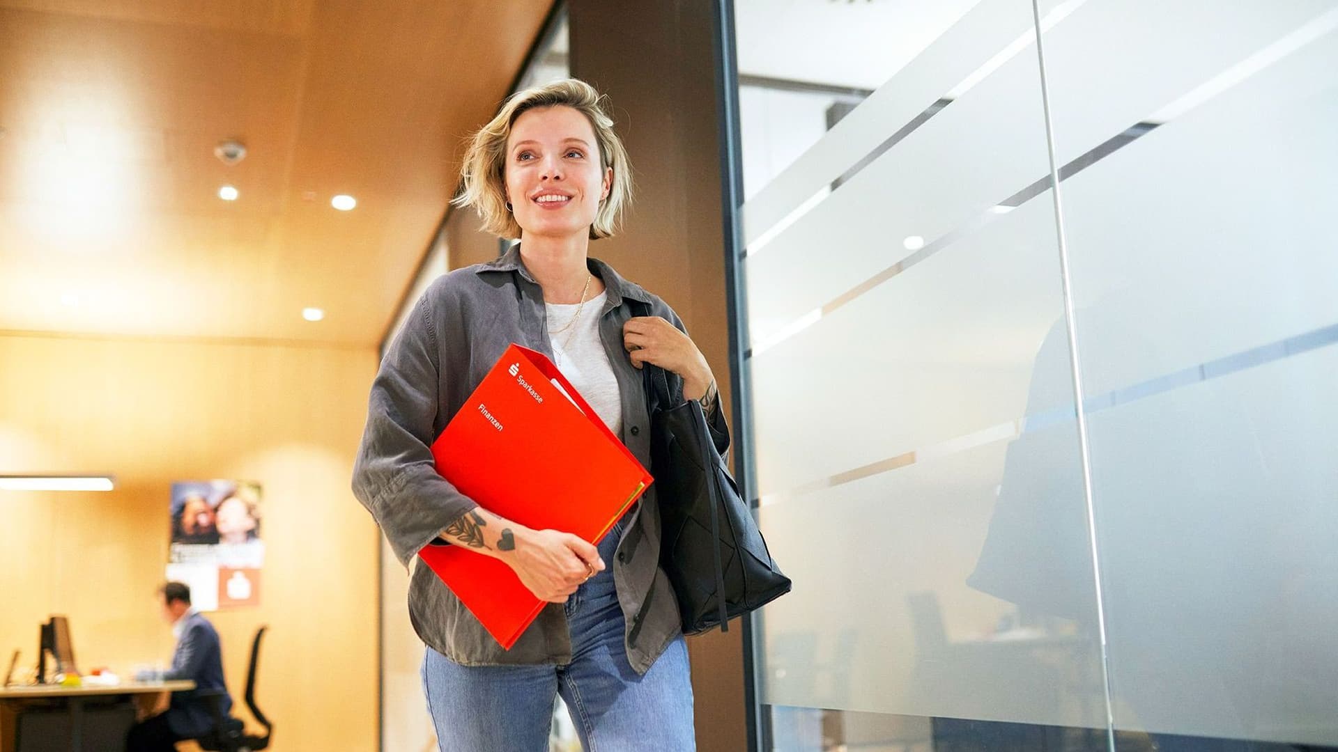Eine junge Frau in einer Sparkassenfinale. Sie läuft lächelnd mit einer roten Mappe unter dem Arm an einer Glaswand vorbei.