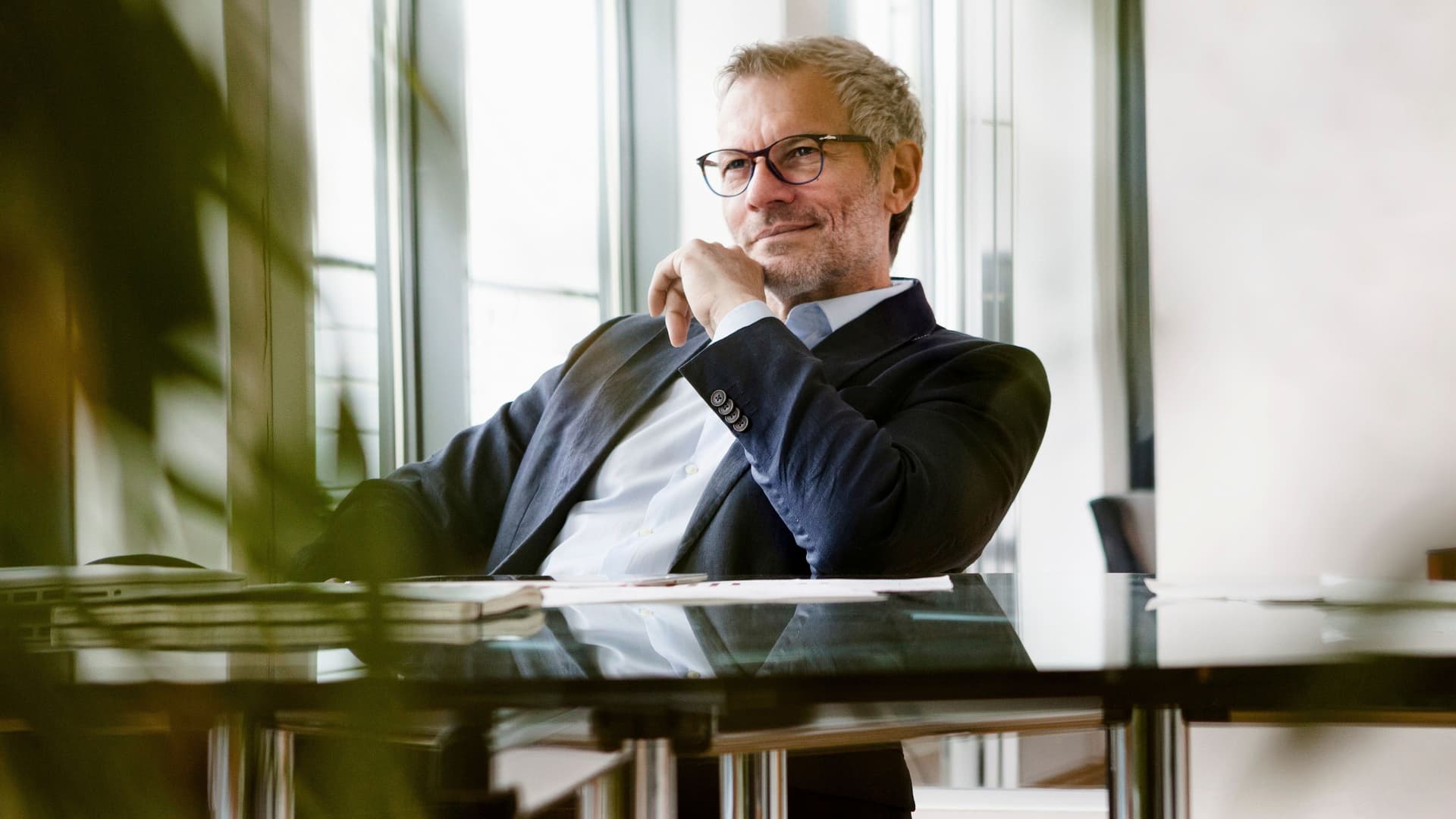 Mann im Anzug sitzt zurückgelehnt an einem Tisch in einem Büro und guckt nach vorne.