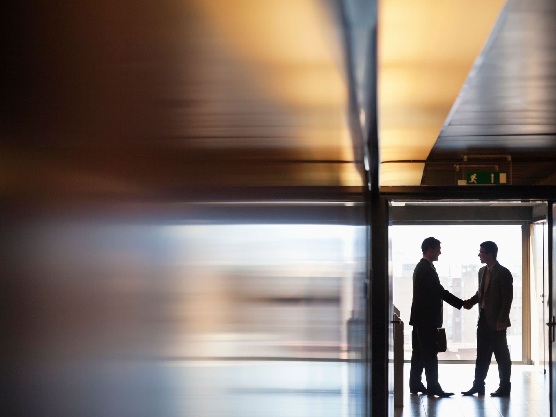 Zwei Männer in Business-Kleidung schütteln sich die Hände in einem Korridor. Es sind nur ihre Silhouetten erkennbar.