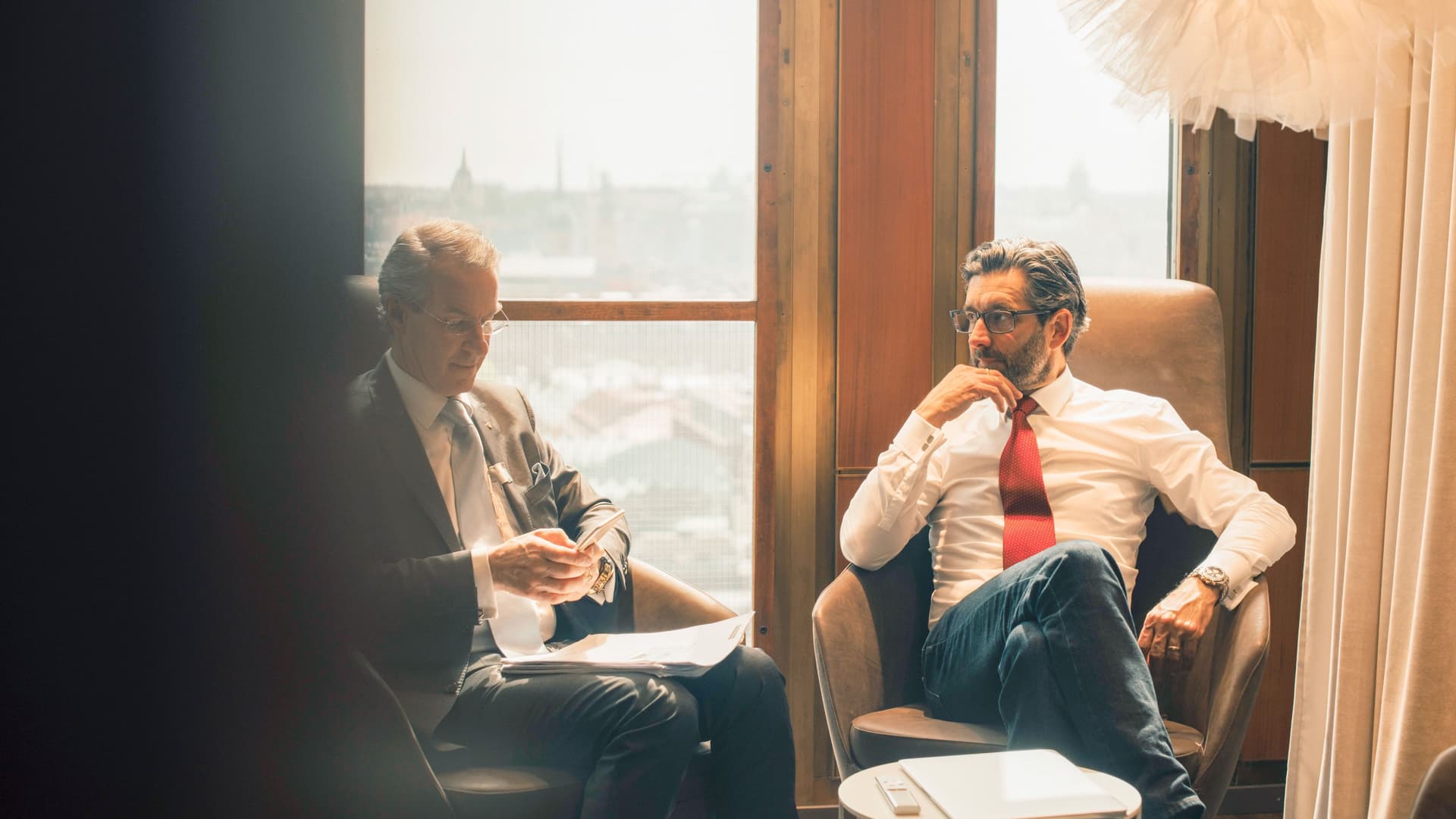 Zwei Männer in Business Kleidung sitzen diskutierend vor einem großen Fenster.