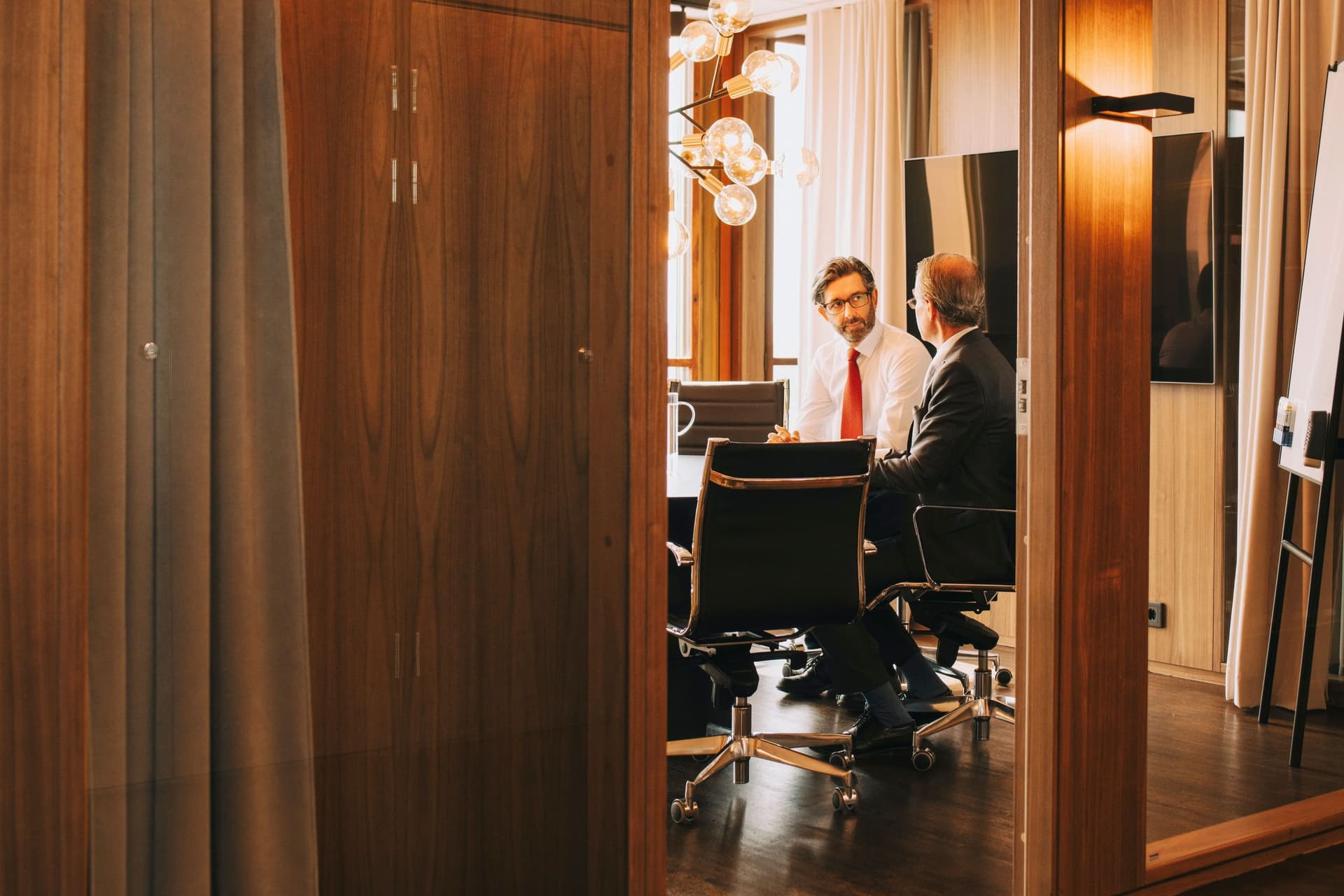 Zwei Männer mittleren Alters sitzen in einem Meeting Raum. Hinter ihnen ist ein großer Bildschirm und ein Whiteboard zu sehen.