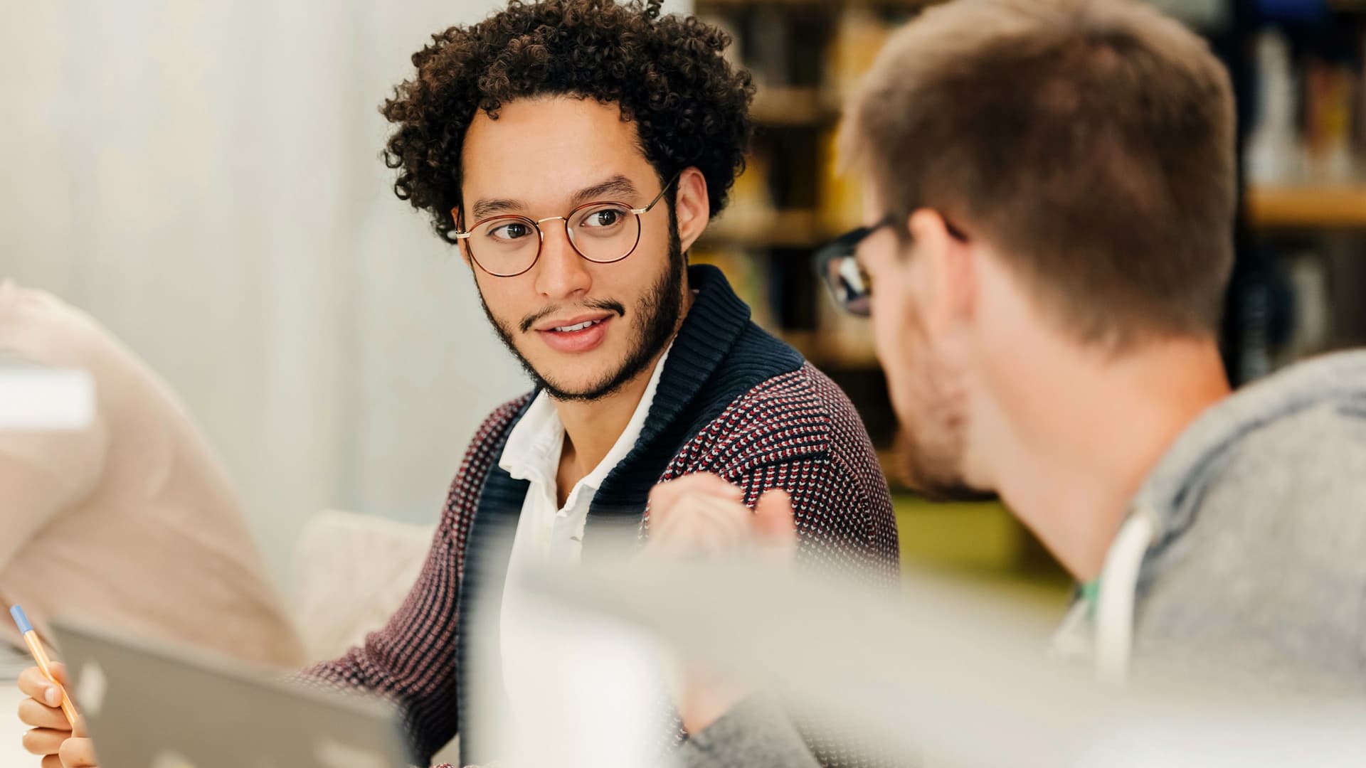 Zwei Studenten vor ihren Laptops, die sich beim Lernen in der Bibliothek leise unterhalten.