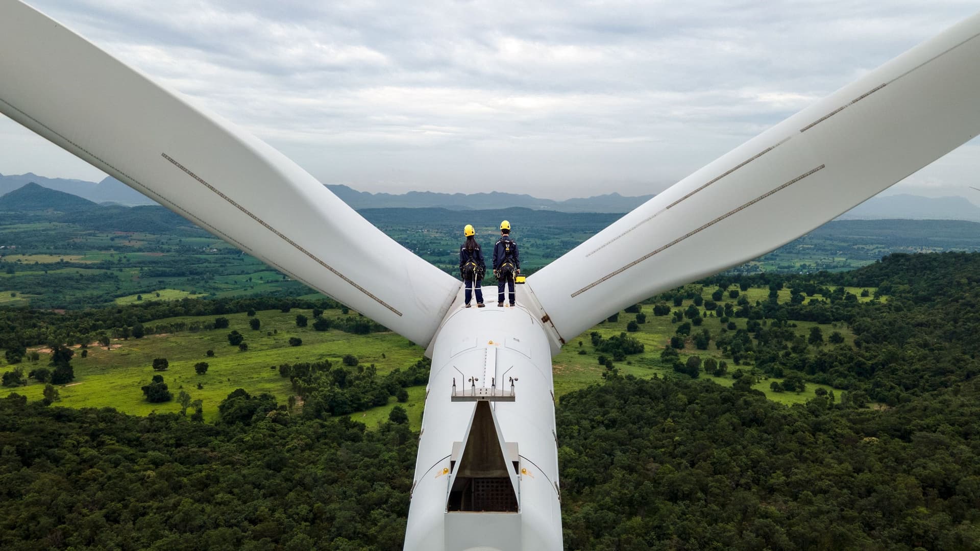 Zwei Elektroingenieure mit Schutzausrüstung stehen oben auf einem Windrad zwischen den Windblättern. Sie schauen in die Landschaft.
